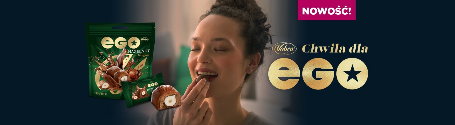 Uśmiechnięta kobieta z zamkniętymi oczami wkłada do ust czekoladową pralinkę. Obok znajduje się zielone opakowanie produktu EGO Hazelnut Passion, przekrojona pralina oraz hasło „NEW! Vobro A moment for EGO” na ciemnym tle.
