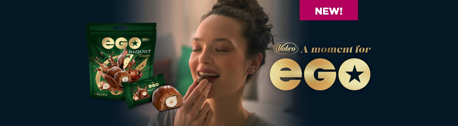 A smiling woman with her eyes closed places a chocolate praline into her mouth. Next to her is the green packaging of the product EGO Hazelnut Passion, a cut-open praline, and the slogan “NEW! Vobro A moment for EGO” set against a dark background.