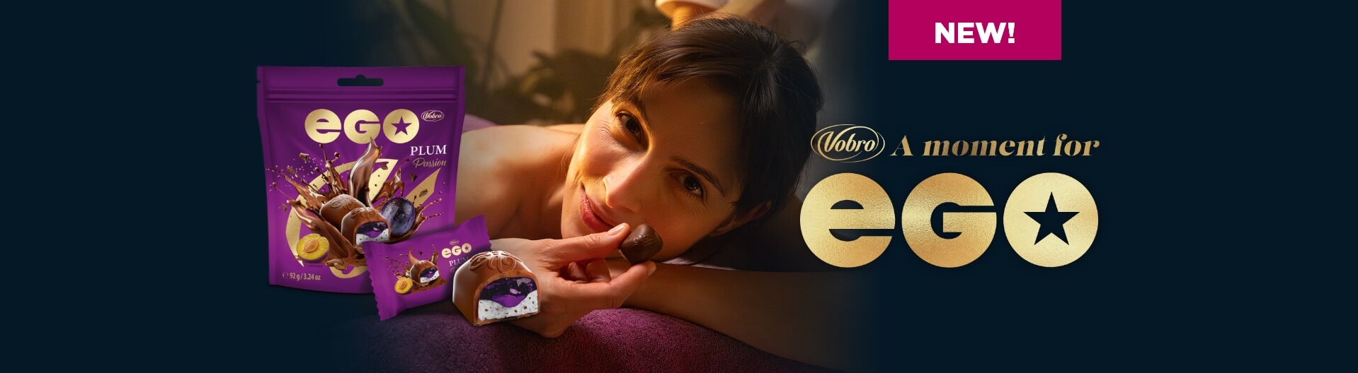 A woman lies on her stomach during a massage, gently smiling as she holds a chocolate praline close to her lips. Next to her, the purple packaging of EGO Plum Passion is visible, along with a cut-open praline revealing plum filling and the slogan “NEW! Vobro. A moment for EGO” set against a dark background.